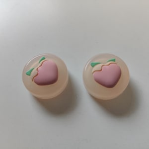 Op de afbeelding: Twee siliconen controller-kappen voor gameconsoles in de vorm van een perzik. De kappen zijn doorschijnend en hebben een roze perzikontwerp met groene bladeren.