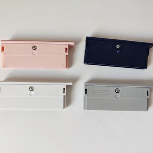 Pode incluir: Quatro cartuchos de Game Boy Advance em rosa pastel, azul marinho, branco e cinza. Cada cartucho tem formato retangular com um pequeno parafuso e o logotipo da Nintendo. Os cartuchos estão dispostos sobre uma superfície branca.