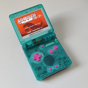 May include: A teal Game Boy Advance SP with a pink button pad and a screen displaying a game title in Japanese. The game title is "Dragon Quest Monsters Joker".