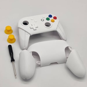 Puede incluir: Mando de juego blanco desmontado con joysticks amarillos y un destornillador negro. El mando tiene botones de colores y una cruceta. Los componentes están dispuestos sobre una superficie blanca, lo que sugiere reparación o personalización.