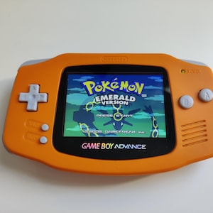 以下が含まれることがあります： 「ポケットモンスター エメラルド」のゲームが表示されたオレンジ色のゲームボーイアドバンス。画面にはゲームタイトルとスタイリッシュなグラフィックが表示されています。コンソールには十字キー、ボタン、そして「Game Boy Advance」のロゴがあります。