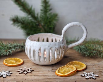 May include: A white ceramic candle holder with a handle, featuring vertical cutouts. The holder sits on a wooden surface with dried orange slices, wooden snowflakes, and evergreen sprigs. A lit candle is inside.