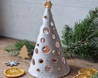 May include: A ceramic Christmas tree candle holder with a gold star on top. The white tree has circular cutouts and gold accents. Decorative wooden ornaments and dried orange slices surround the tree on a wooden surface.