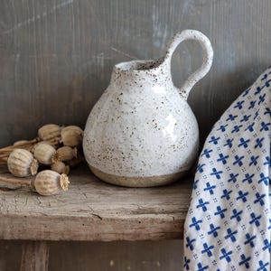 Może przedstawiać: Biały dzbanek ceramiczny z kropkowanym szkliwem i zaokrąglonym korpusem. Dzbanek ma pojedynczy uchwyt i lekko rozszerzoną krawędź. Stoi na rustykalnym drewnianym stołku.