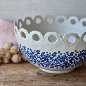 Może przedstawiać: Biała ceramiczna miska z falistym brzegiem i niebieskim wzorem kwiatowym. Misa stoi na drewnianej powierzchni. Obręcz ma równomiernie rozmieszczone okrągłe wycięcia. Niebieski wzór to powtarzający się wzór małych liści i kropek.