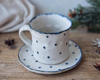 May include: A white ceramic mug with a blue rim and blue polka dots. The mug is sitting on a matching saucer with a blue rim and blue polka dots. The mug and saucer are on a wooden surface.