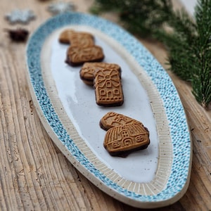Könnte beinhalten: Eine weiße ovale Keramik-Servierplatte mit einem blau-weißen gesprenkelten Design. Die Platte ist mit Lebkuchen in Form von Häusern und Windmühlen gefüllt.