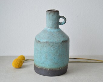 May include: A small, turquoise ceramic vase with a brown glaze and a handle. The vase has a rounded body and a narrow neck.
