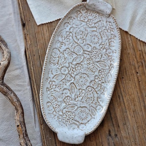 Könnte beinhalten: Ein ovales Serviertablett aus Keramik mit weiß glasierter, gesprenkelter Oberfläche und einem komplexen, geprägten Blumenmuster. Das Tablett verfügt über zwei kleine Griffe an den gegenüberliegenden Enden und einen strukturierten Perlenrand.