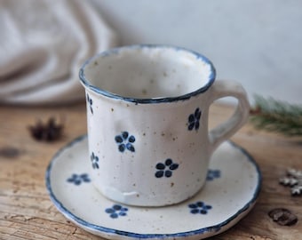 May include: A white ceramic coffee cup with a blue rim and blue floral pattern, sitting on a matching saucer. The cup and saucer are on a wooden surface.