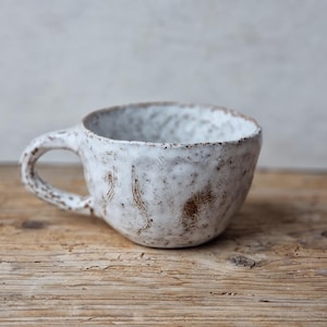 Op de afbeelding: Een witte keramische mok met een textuur, gespikkeld oppervlak en een afgeronde handgreep. De mok staat op een houten oppervlak.