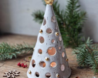 May include: A white ceramic Christmas tree-shaped candle holder with a gold star on top. The tree has circular cutouts and gold accents. Decorative wooden snowflakes, a small wooden tree, and red berries are scattered around the base.