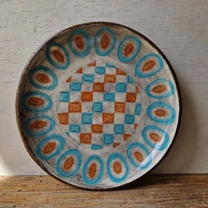 May include: A ceramic plate with a white background and a geometric design in shades of blue and orange. The design features a checkerboard pattern in the centre surrounded by a ring of oval shapes.