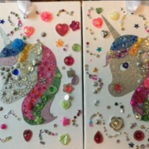 May include: Two rectangular white panels featuring glittery unicorn silhouettes decorated with an assortment of colourful plastic gems, beads, and sequins in various shapes including hearts, stars, and flowers, with white ribbons attached at the top.