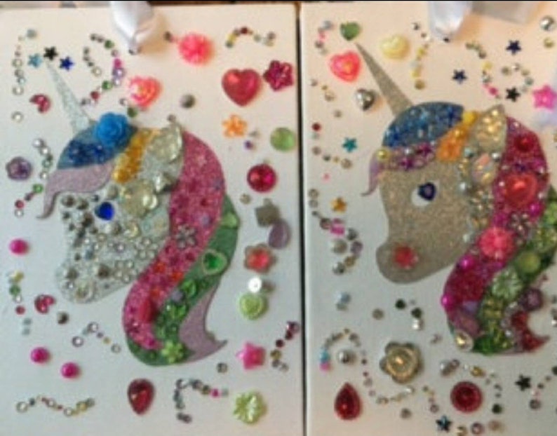 May include: Two rectangular white panels featuring glittery unicorn silhouettes decorated with an assortment of colourful plastic gems, beads, and sequins in various shapes including hearts, stars, and flowers, with white ribbons attached at the top.