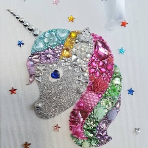 May include: A decorative unicorn wall hanging featuring a silver glitter face with a blue heart-shaped eye. The mane is composed of various colourful beads, rhinestones, and floral-shaped embellishments in pink, green, and purple. Small colourful stars are scattered around the unicorn.