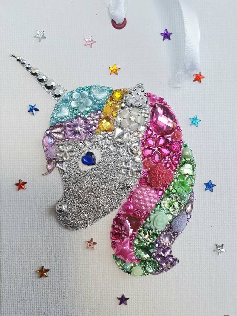 May include: A decorative unicorn wall hanging featuring a silver glitter face with a blue heart-shaped eye. The mane is composed of various colourful beads, rhinestones, and floral-shaped embellishments in pink, green, and purple. Small colourful stars are scattered around the unicorn.