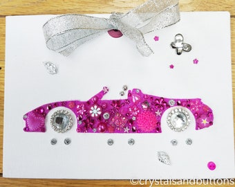 Puede incluir: Una tarjeta rectangular blanca con la silueta de un coche descapotable rosa adornada con varios brillantes, cuentas y botones florales rosas y transparentes. Un lazo plateado está fijado en la parte superior, con el texto crystalsandbuttons abajo.