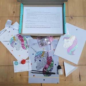 May include: A craft kit featuring three unicorn-themed art projects, a box of instructions, a small pink mesh bag of assorted embellishments, black tweezers, a small glue bottle, a white triangular tray, a wooden stick, and a gift tag.