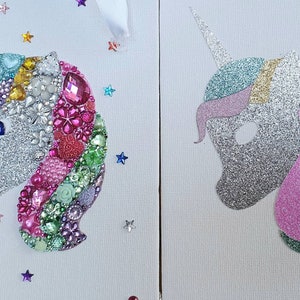 May include: Two side-by-side unicorn head wall art pieces on white canvas. The left unicorn is decorated with multicoloured rhinestones, gems, and floral embellishments. The right unicorn features a silver, pink, and green glitter finish.