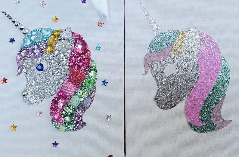 May include: Two side-by-side unicorn head wall art pieces on white canvas. The left unicorn is decorated with multicoloured rhinestones, gems, and floral embellishments. The right unicorn features a silver, pink, and green glitter finish.
