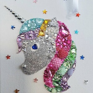 May include: A decorative unicorn wall hanging featuring a silver glitter face with a blue heart-shaped eye. The mane is composed of various colourful beads, rhinestones, and floral-shaped embellishments in pink, green, and purple. Small colourful stars are scattered around the unicorn.