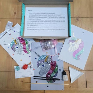 May include: A craft kit featuring three unicorn-themed art projects, a box of instructions, a small pink mesh bag of assorted embellishments, black tweezers, a small glue bottle, a white triangular tray, a wooden stick, and a gift tag.