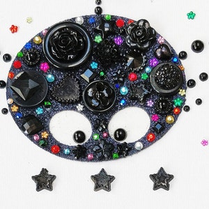 Può includere: Una base circolare glitterata blu scuro decorata con un assortimento di bottoni neri, tra cui forme di rose, cuori e stelle, intervallati da piccoli strass e perline colorate. Due grandi aperture circolari fungono da occhi.