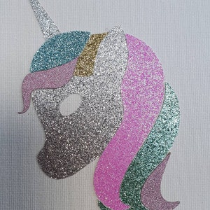 May include: A glittery unicorn head cutout featuring a silver face and horn, a teal and gold forehead section, and a flowing mane composed of layered pink, teal, and silver glitter segments. The design is flat and oriented in profile.