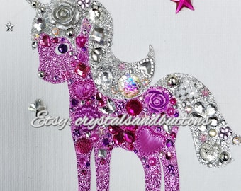 May include: A decorative unicorn applique featuring a glittery pink body and a shimmering silver mane and tail. The design is embellished with various pink and clear crystals, buttons, and floral accents. The text Etsy: crystalsandbuttons is visible.