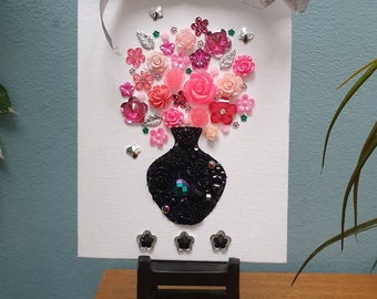 May include: A white rectangular canvas featuring a collage of pink and red floral embellishments arranged above a black vase made of beads and sequins. A silver ribbon is tied at the top, and three small black flower-shaped charms are placed below the vase.