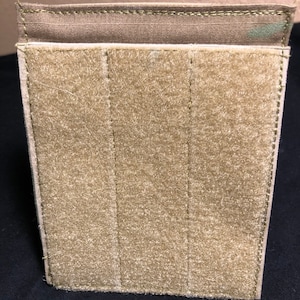 May include: A tan rectangular pouch with a hook and loop fastener surface. The pouch has a folded top edge and vertical stitched lines. The background is a dark surface.