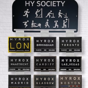 May include: A black metal display rack with the text HY SOCIETY and white icons of fitness exercises. Below are three vertical columns of rectangular fabric patches, each featuring the text HYROX and various city names like LON, BIRMINGHAM, TORONTO, MAASTRICHT, CARDIFF, LAS_VEGAS, MADRID, GLASGOW, and BOSTON.