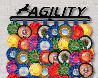 Puede incluir: Un soporte de metal negro para premios de agility para perros con la palabra "AGILITY" en letras grandes. El soporte tiene múltiples ganchos para colgar cintas. Los ganchos están llenos de cintas de colores de concursos caninos con diferentes posiciones, incluyendo "1º", "2º", "3º", "4º", "6º", "Ronda limpia" y "Mejor de raza".