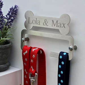 May include: A silver metal dog leash holder shaped like a bone with the names "Lola & Max" engraved on it. Three leashes are hanging from the holder, one red with white paw prints, one red with white bone prints, and one blue with white polka dots.