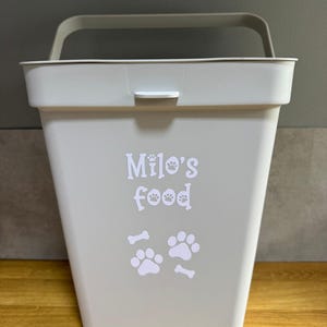 May include: A pale grey pet food storage container with a handle and a latch. The container is labelled "Milo's food" in white, with paw prints and bone graphics.