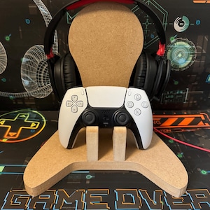 May include: A wooden stand with a cutout in the shape of a controller, designed to hold a white wireless controller and a black headset with red accents. The stand is sitting on a black and white surface with the text "GAME OVER" printed on it.