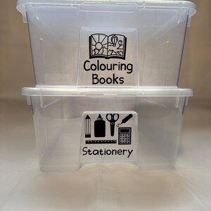 May include: Two clear plastic storage boxes, one on top of the other. The top box has a label reading "Colouring Books" with a mountain scene graphic. The bottom box has a "Stationery" label with school supply icons.