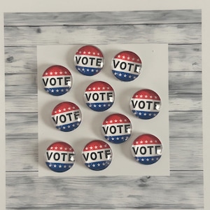 May include: A collection of ten round buttons with a red, white, and blue design. Each button has the word "VOTE" in white text. The buttons are arranged on a white surface with a light gray wood-grain background.