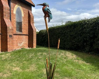 May include: A metal sculpture of a kingfisher bird perched on cattails. The bird has blue and orange coloring. The cattails and reeds are made of metal and are brown and green. The sculpture is placed in a grassy yard.