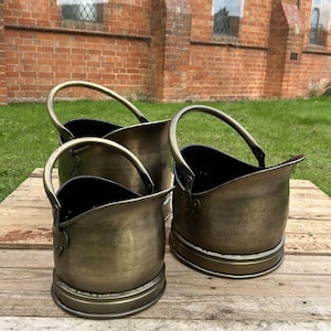 May include: Three antique brass coal buckets with arched handles. The buckets have a rounded base and a dark interior. They are arranged on a wooden surface with a brick building in the background.