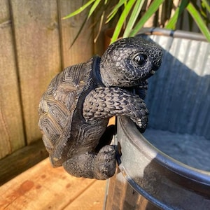 May include: Black turtle statue with a textured shell, climbing the side of a metal planter. The turtle's head is turned to the side, and its eyes are closed.