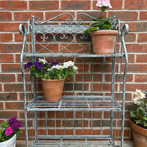 May include: A grey, three-tiered metal plant stand with decorative scrollwork and heart-shaped details. It displays terracotta pots filled with colourful flowers, including pink, purple, and white. The stand is positioned in front of a brick wall.