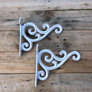 May include: Two matching wall brackets featuring a white distressed finish over a dark base. Each bracket has a vertical mounting plate with screw holes and a curved, scrollwork support arm extending outward in a decorative pattern.