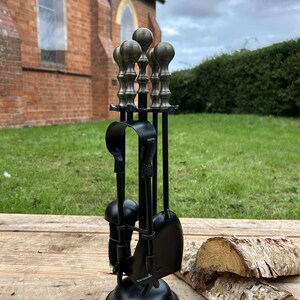 May include: A black fireplace tool set with a round base and a set of tools, including a poker, shovel, and brush. The handles of the tools have a decorative, antique brass finish. The set is displayed on a wooden surface with logs in the background.