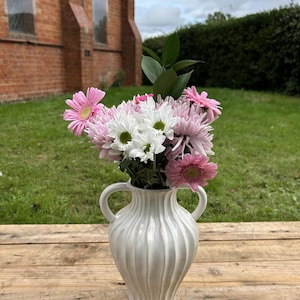 May include: A white ceramic vase with a ribbed design, holding a bouquet of pink and white flowers. The vase has two handles and sits on a wooden surface. The flowers include pink gerbera daisies and white chrysanthemums.
