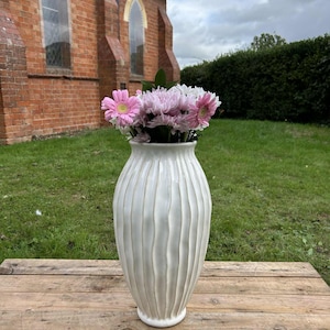 May include: A cream-coloured ceramic vase with vertical ribbed detailing, holding pink and white flowers. The vase is on a weathered wooden surface, with a brick building and green lawn in the background.