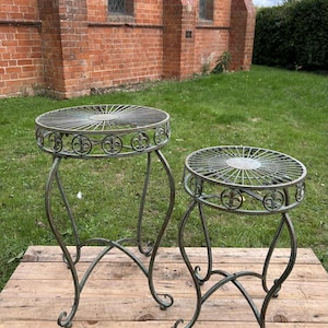 May include: Two antique metal plant stands. The stands have a circular top with a decorative fleur-de-lis design around the edge. The legs are curved and ornate. The stands are on a wooden surface and are set against a brick building and green grass.
