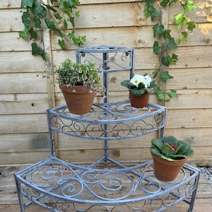 May include: A three-tiered, grey metal plant stand with a decorative scroll design. It holds three terracotta pots with flowering plants. Green ivy climbs the wooden fence in the background.