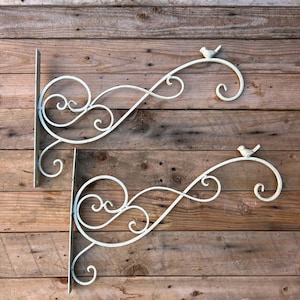 May include: Two white metal wall brackets featuring scrollwork designs and a small bird figure perched on the top curve of each arm. The brackets have a flat vertical mounting plate with pre-drilled holes for attachment to a wall surface.